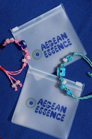 Two woven adjustable rope evil eye bracelets on clear 'Aegean Essence' packaging against a blue background