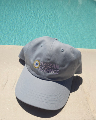 Light blue baseball hat (dad hat) with embroidered evil eye and Aegean Essence logo in front of a swimming pool