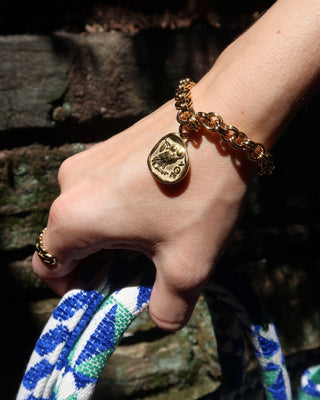 A close-up of a person's hand wearing a gold chain bracelet with a Greek Athena goddess of wisdom owl coin pendant and a matching gold ring, set against a dark, rustic background with a blue and white patterned textile visible at the bottom.