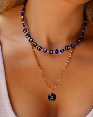 Woman wearing a gold chain necklace with a round dark blue evil eye pendant and a blue glass evil eye choker necklace layered. Greek jewelry made in Greece