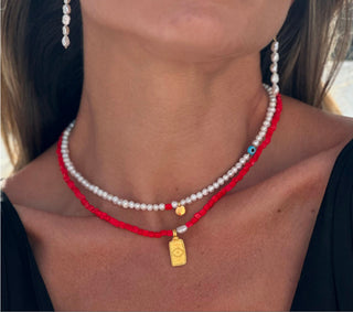 Model wearing a freshwater pearl necklace with an evil eye and a red beaded necklace with a gold evil eye charm made in Greece. Greek jewelry at Aegean Essence