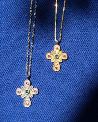 Two ornate cross-shaped pendant necklaces displayed on a textured blue fabric background. One necklace has a silver-toned chain with a silver cross pendant featuring a green emerald center stone and red ruby accent stones.
