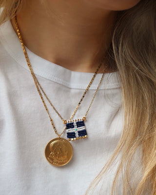 Woman wearing a gold Poseidon ancient greek god coin necklace and  a woven macrame Greek flag necklace with a gold chain and gold bead details made in Greece. Vintage Hellenic Republic flag necklace with turquoise stone and gold embellishments