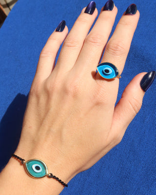 Woman's hand and wrist wearing a turquoise blue Murano glass 18k gold plated evil eye bracelet with an adjustable black woven design made in Greece, Greek evil eye jewelry and a blue glass evil eye ring