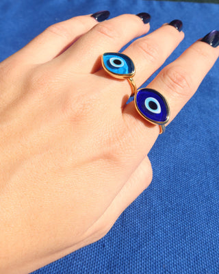 Hand wearing a light blue Murano glass evil eye ring with a gold band and a dark blue Murano glass evil eye ring with a gold band against a blue background