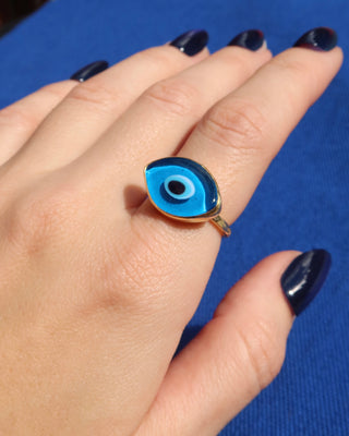 Woman's hand wearing a light blue Murano glass evil eye ring with a gold band against a blue background, Greek evil eye jewelry