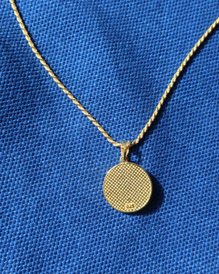 A gold necklace with a textured circular pendant on a blue woven fabric background.