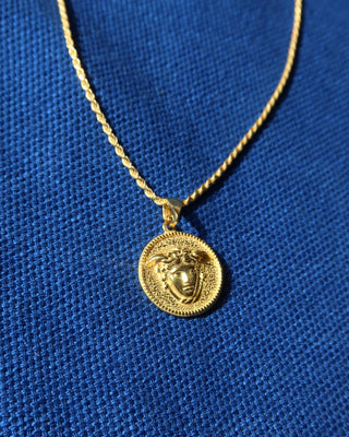 A gold-tone chain necklace with a circular pendant featuring a Medusa-style head motif, displayed on a textured blue fabric background. Greek mythology jewelry