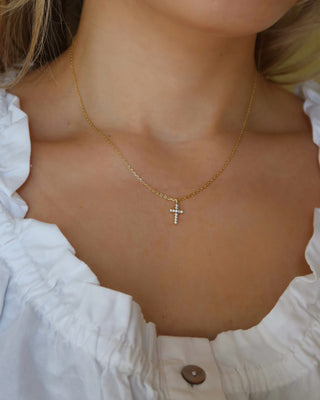 Gold necklace with a cubic zirconia cross pendant worn by a person in a white blouse.