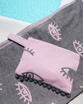 Gray towel with eye pattern and pink pouch with dark gray evil eye print and gray pom pom trim next to a swimming pool