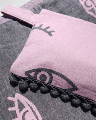 Gray towel with eye pattern and pink pouch with dark gray evil eye print and gray pom pom trim next to a swimming pool