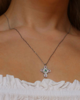 A close-up of a woman's neck and upper chest wearing a delicate silver-toned cross pendant necklace with small ruby and green emerald gemstones, paired with a white ruffled top.