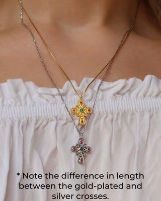 Two layered cross necklaces worn on a model's neck over a white off-the-shoulder top. One necklace is gold-plated with a gold cross pendant with emerald and ruby stones, and the other is a longer silver 925 cross necklace of the same design but longer length chain