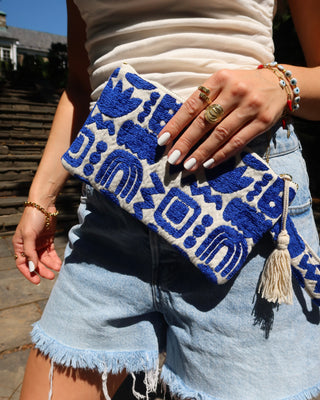 A person outdoors holding a woven clutch bag with a bold blue and off-white geometric pattern, paired with light denim shorts and gold jewelry.