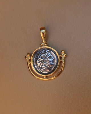 A gold pendant featuring an antique-style circular coin medallion with a raised classical bust of Alexander the Great, displayed on a textured brown background. 18k gold-plated sterling silver 925 made in Greece, Greek coin jewelry