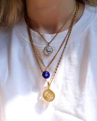Woman wearing a necklace stack with an ancient Greek Heracles coin necklace, a blue glass evil eye necklace on a gold chain, and a sun coin pendant necklace on a long gold chain. Greek jewelry made in Greece