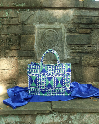 A structured tote bag karge handbag with a blue, green, and white geometric pattern, displayed on a stone ledge with a draped blue satin cloth in front of a rustic stone wall.