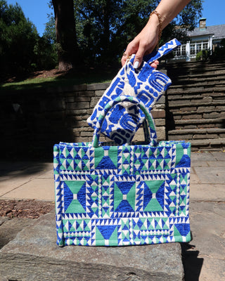 Hand holding a blue and cream wristlet clutch bag with symbols above a green and blue geometric print woven tote bag handbag