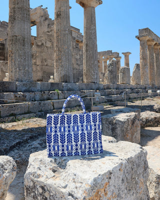 A blue and white patterned evil eye and olive lead woven tote bag with a woven handle sits on a large stone block in front of ancient stone columns and ruins under a clear blue sky in Greece. Emily in Paris handbag 