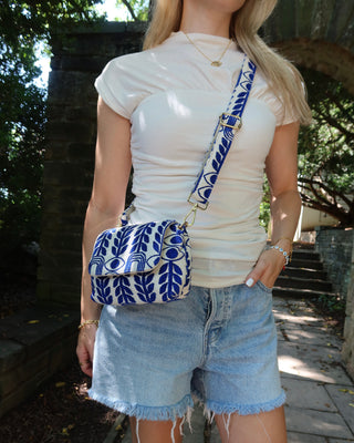 A woman wearing a white ribbed t-shirt and denim shorts carries a blue and white patterned fabric crossbody bag with gold hardware, standing outdoors against a dark brick wall. The bag has a royal blue evil eye and olive branch print made in Greece by the same brand as Emily in Paris Lily Collins handbag