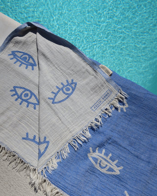 Blue cotton towel with white evil eye patterns  and a reversible white and blue evil eye print on a poolside