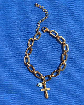 Gold chain link bracelet with a cross charm and a white evil eye charm on a blue background with an extender and lobster clasp