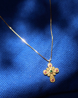 A gold Byzantine style cross pendant necklace with small rubies and emerald stones, resting on a textured blue fabric background.