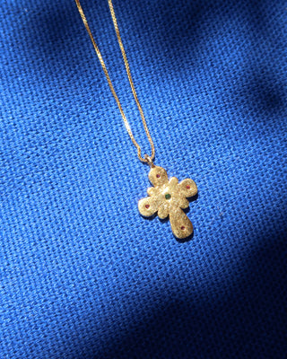 The back of a gold Byzantine style cross pendant necklace with small rubies and emerald stones on a blue background, made in Greece. Greek cross necklace