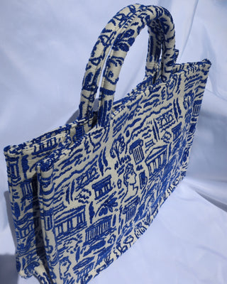 large cream/off-white handbag tote bag with a woven blue Greece print with the Parthenon, ancient Greek sculptures, owls, ionian columns, olive branches, olive trees, and more. Handcrafted bag made in Greece