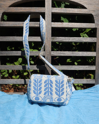 A small fabric crossbody bag with a blue and white evil eye and olive branch pattern, resting on a light blue blanket in front of a weathered wooden fence with green foliage behind it. Made in Greece