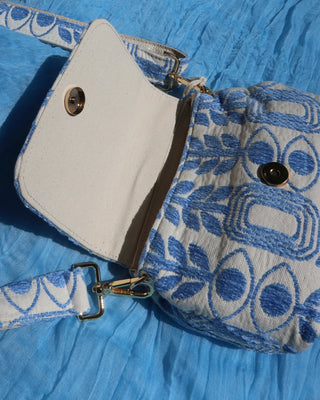 A close-up of a small crossbody bag with a blue and white patterned fabric body, a white canvas flap, and a matching patterned strap, laid on a blue textured fabric background. With gold hardware including a snap closure