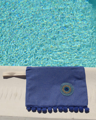 Woven blue clutch bag zipper wet/dry pouch with an embroidered metallic evil eye and a blue pom pom trim next to a pool