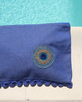 Woven blue clutch bag zipper wet/dry pouch with an embroidered metallic evil eye and a blue pom pom trim next to a pool