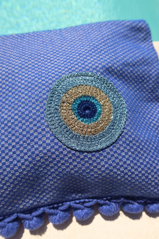 Woven blue clutch bag zipper wet/dry pouch with a circular embroidered metallic evil eye and a blue pom pom trim next to a pool