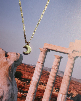 Gold necklace made in Greece with a crescent moon pendant against a scenic background of ancient Greek stone columns and landscape.
