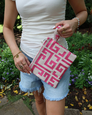 A woman outdoors holding a pink and white Greek key meander print clutch bag. She is wearing a white tank top, denim shorts, and gold jewelry, with green garden foliage in the background.