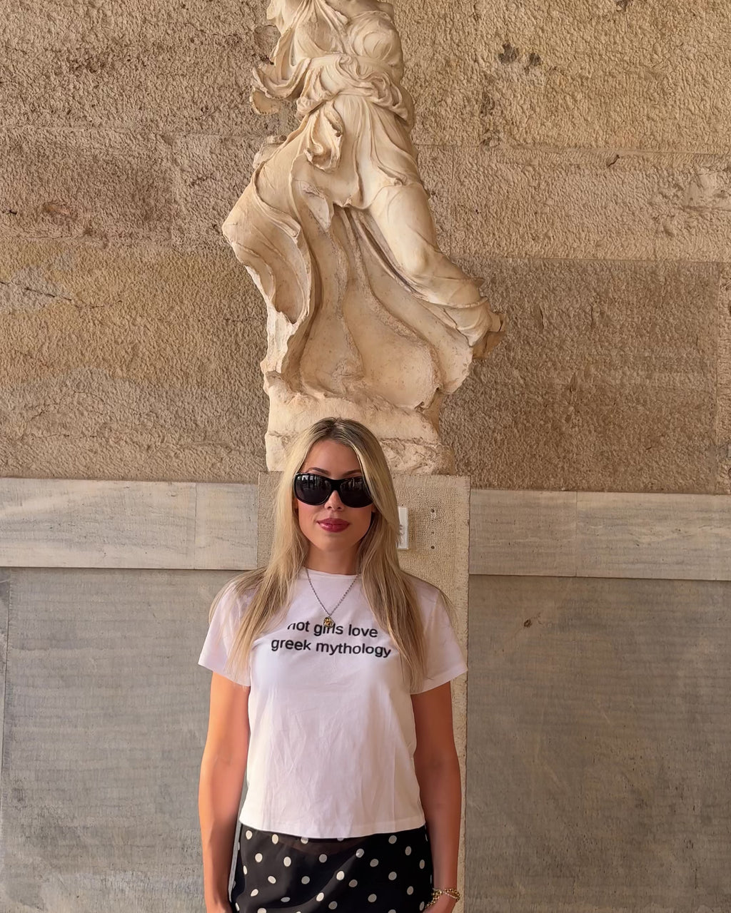 Woman wearing a "hot girls love greek mythology" graphic print t-shirt with a black and white polka dot print midi skirt standing in front of an ancient greek statue