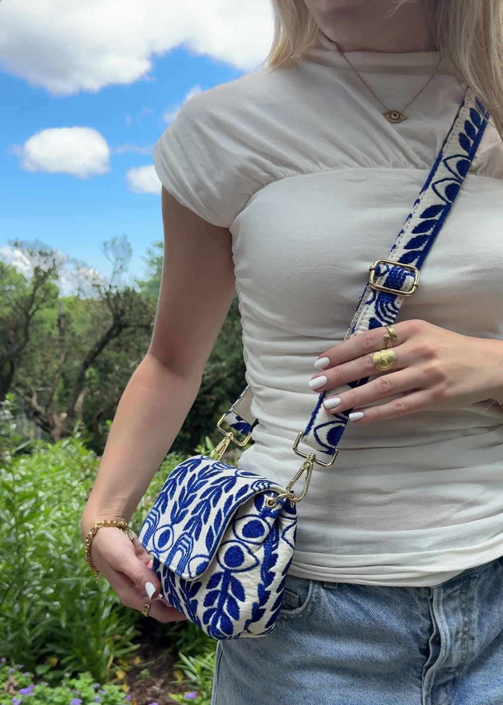 A woman wearing a white ribbed t-shirt and denim shorts carries a blue and white patterned fabric crossbody bag with gold hardware, standing outdoors against a dark brick wall. The bag has a royal blue evil eye and olive branch print made in Greece by the same brand as Emily in Paris Lily Collins handbag