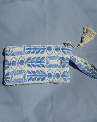 A small woven clutch or pouch with a blue and off-white geometric pattern with evil eye print and olive branches, featuring a wrist strap and a cream tassel zipper pull, laid flat on a blue fabric background. Marabu Greek brand at Aegean Essence