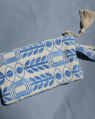 A small woven clutch or pouch with a blue and off-white geometric pattern with evil eye print and olive branches, featuring a wrist strap and a cream tassel zipper pull, laid flat on a blue fabric background. Marabu Greek brand at Aegean Essence