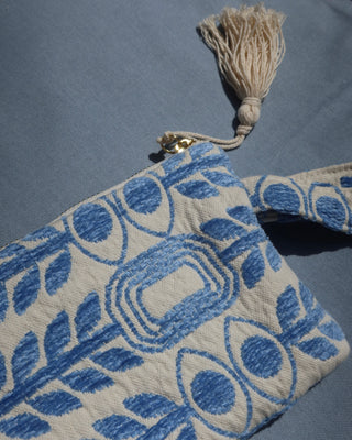 A close-up of a woven fabric pouch or clutch with a blue and cream evil eye and olive branch pattern, featuring a cream tassel and a small gold-tone metal clasp, laid on a blue textured background. Marabu Greek brand at Aegean Essence