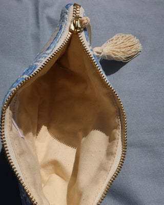 An open blue and white patterned clutch or pouch with a gold zipper, cream fabric lining, and a cream tassel, shown on a blue-gray fabric background with a cotton canvas interior